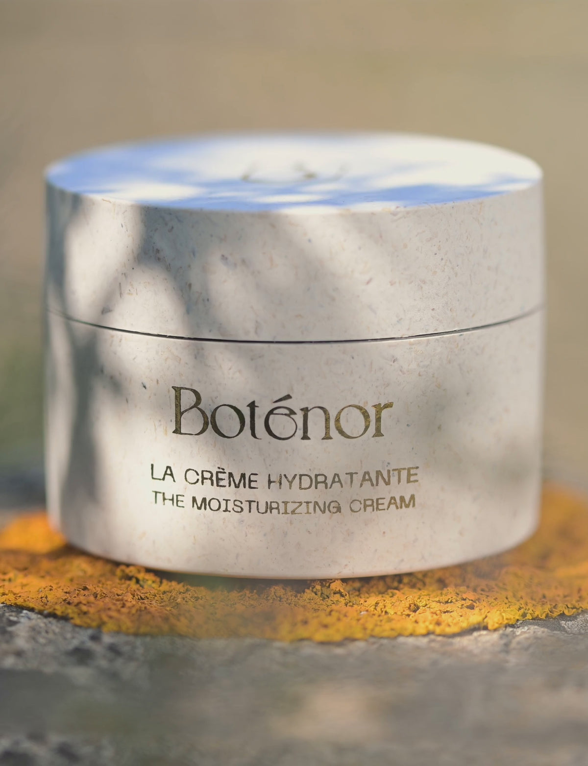 Gros plan du pot de La Crème Hydratante Boténor, posé sur un rond de lichen orangé, à l'ombre d'oliviers sauvages